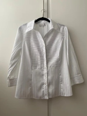 Vintage vit  långärmad skjorta med subtila ränder - Vit skjorta med subtila vertikala ränder och klassisk krage. Skjortan har långa ärmar med breda manschetter och är tillverkad i ett glansigt material som ger en elegant känsla. Perfekt för dig som gillar stilrena plagg med lite extra detalj.