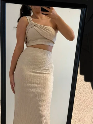 Beige ribbat set med crop top - Säljer ett beige ribbat set med en asymmetrisk crop top med bred axelband och twistad detalj framtill. Kjolen är högmidjad och lång med slits på sidan. Materialet är stretchigt och mjukt, perfekt för en trendig look.