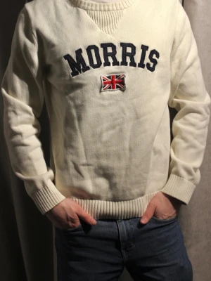 Vit stickad tröja från Morris - ‼️Står XL men passar som M‼️.                         Vit stickad tröja från Morris med mörkblå text och en broderad brittisk flagga på bröstet. Tröjan har ribbade muddar vid ärmslut och nederkant samt en klassisk rund halsringning med ribbad detalj. Perfekt för en stilren och avslappnad look. Modellen är 182 cm lång 