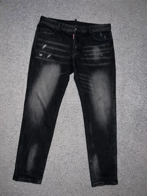 Svarta jeans från Dsquared2 - Coola svarta jeans från Dsquared2 med slitna detaljer och ljusa partier. Modellen har raka ben, klassiska fyra fickor och en tydlig 'CIAO' patch på bakfickan. Perfekt för dig som gillar en edgy och trendig look.