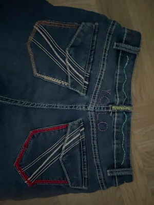 Blå YOLO jeans med färgglada sömmar - Säljer ett par blå jeans med unika färgglada sömmar på bakfickorna i rött, beige och vitt. Jeansen har bootcut-modell och detaljerade fickor som sticker ut. Perfekta för dig som vill ha något annorlunda och coolt i garderoben.