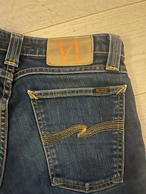 Blå jeans från Nudie Jeans - Säljer ett par klassiska blå jeans från Nudie Jeans med snygga kontrastsömmar och ikonisk brodyr på bakfickan. Jensen har raka ben och en ganska låg midja.
