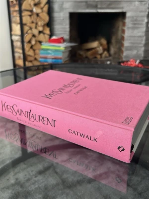 Yves Saint Laurent Catwalk bok - Säljer en snygg och inspirerande bok 'Yves Saint Laurent Catwalk' med rosa hård pärm. Boken innehåller haute couture-kollektioner och är perfekt för modeintresserade. Tryckt av Thames & Hudson, med stilrena fotografier och texter om designern.
