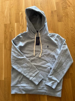 Grå hoodie från Polo Ralph Lauren - Snygg grå hoodie från Polo Ralph Lauren med klassisk huva och vita dragsnören. Tröjan har en broderad logga på bröstet och en stor magficka framtill. Tillverkad i mjukt bomullsmaterial som känns skönt mot huden. Perfekt för en avslappnad och stilren look.