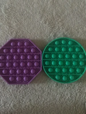 Två pop it fidget-leksaker - Säljer två pop it fidget-leksaker i silikon, en lila med åttakantig form och en grön som är rund. Perfekta för att hålla händerna sysselsatta och minska stress. Båda har bubblor att trycka på och är lätta att ta med sig.