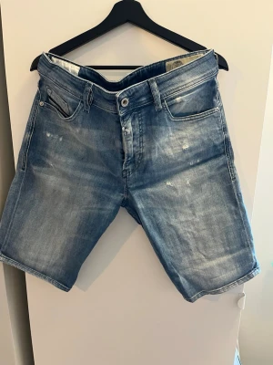 Blå jeansshorts från Diesel - Säljer ett par blå jeansshorts från Diesel med slitna detaljer och klassisk femficksdesign. Shortsen har normal passform och är tillverkade i bomull. Perfekt för dig som gillar en avslappnad och cool stil. Storlek 31 