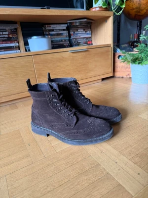 Bruna mocka brogue-stövletter - Snygga bruna stövletter i mocka med klassiska broguedetaljer och snörning framtill. Skorna har robust sula och dragflik bak för enkel påtagning. Perfekta för dig som gillar en stilren och tidlös look.