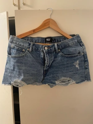 Blå jeanskjol från 157 med slitningar - Säljer en blå jeanskjol från 157 med rå kant och slitna detaljer framtill. Kjolen har klassisk femficksmodell, bälteshällor och dragkedja med knapp. Perfekt för dig som gillar en avslappnad och trendig stil.