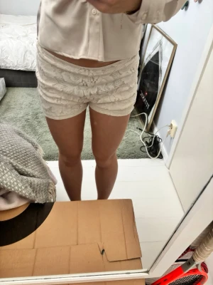 Vita spetsade shorts med dragkedja - Säljer ett par begie vita shorts med blommigt spetsmönster och dragkedja bak. De är mid waist och har en tight passform som framhäver siluetten. Perfekta för sommarens varma dagar och snygga att matcha med en enkel topp.