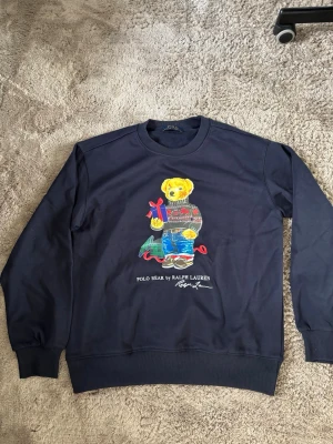 Mörkblå sweatshirt Polo Bear - Mörkblå sweatshirt från Polo Ralph Lauren med ikonisk Polo Bear-motiv på framsidan. Björnen har en julig stickad tröja och håller ett presentpaket. Rund halsringning och ribbade muddar. Perfekt för dig som gillar klassiska och lekfulla plagg.