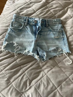 Ljusa jeansshorts från Pull&Bear - Säljer ett par ljusblå jeansshorts från Pull&Bear med råa, fransiga kanter och slitningar framtill. Klassisk femficksmodell med bälteshällor och dragkedja. Perfekta för en avslappnad och trendig sommarstil.