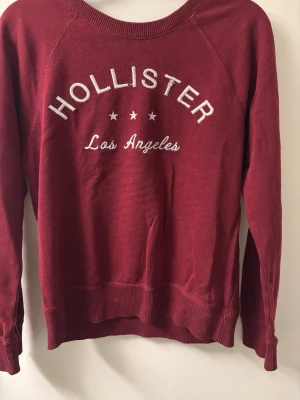 Vinröd sweatshirt från Hollister - Vinröd sweatshirt från Hollister med vit broderad text 'Hollister Los Angeles' och tre stjärnor på bröstet. Tröjan har rund halsringning, långa ärmar och ribbade muddar. Perfekt för en avslappnad och cool look.