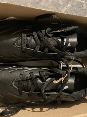 Adidas svarta fotbollsskor - Svarta fotbollsskor från Adidas med vita detaljer och klassisk låg profil. Skorna har snörning och är tillverkade i syntetmaterial som ger bra komfort och grepp på planen. Perfekta för dig som vill ha stilrena och funktionella skor till matchen.