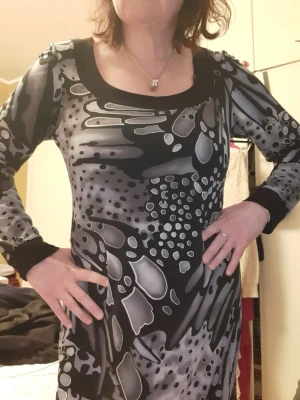 Svart och grå mönstrad klänning - Säljer en svart och grå kortklänning med abstrakt mönster. Klänningen har långa ärmar, bred svart kant vid halsringning och ärmslut samt en rak passform. Perfekt för dig som gillar unika prints och vill sticka ut lite extra. Storlek L. Mått på bilderna. 