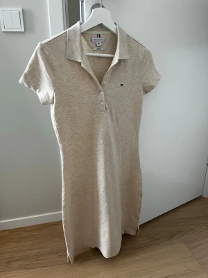 Beige pikéklänning från Tommy Hilfiger - Snygg beige pikéklänning från Tommy Hilfiger med klassisk krage, korta ärmar och knappar framtill. Klänningen har en liten logga på bröstet och är gjord i mjuk bomull, perfekt för en avslappnad och stilren look. Den är i väldigt bra skick och är bara använd en gång😊💖🌷