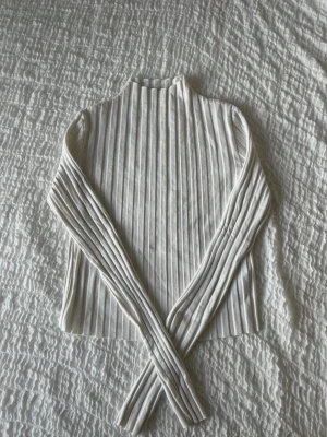 Vit ribbstickad polotröja - Säljer en vit ribbstickad polotröja med lång ärm och hög hals. Tröjan har en tight passform och är gjord i ett mjukt, stretchigt material som känns skönt mot huden. Perfekt att styla med jeans eller kjol för en clean look.