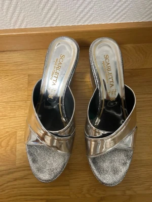 Silvriga platåpumps från Scarletta - Säljer ett par glittriga silvriga platåpumps från Scarletta. Skorna har korslagda band i metallic och en hög kilklack i samma silvriga ton. Perfekta för dig som vill sticka ut med en riktigt snygg och trendig look.