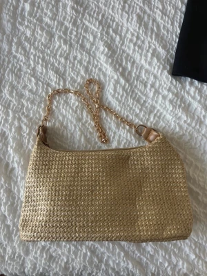 Beige axelväska med kedjerem - Snygg beige handväska i flätad strådesign med guldfärgad kedjerem. Väskan har en enkel, rektangulär form och stängs med dragkedja. Perfekt för dig som gillar en trendig och naturlig look.