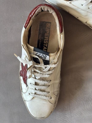 GGDB Superstar sneakers med röd stjärna - Snygga GGDB Superstar sneakers i vitt läder med en röd stjärna på sidan och röda detaljer vid hälen. Klassisk låg modell med vita snören och vintage-inspirerad design. Perfekta för dig som gillar en cool och avslappnad stil.