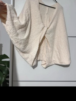 Krämvit fluffig kofta/poncho aktig - Mysig och fluffig kofta i krämvit färg med knut framtill. Koftan har vida ärmar och är tillverkad i ett mjukt, stickat material som känns riktigt skönt mot huden. Perfekt för att slänga över en topp när det är lite kyligt.