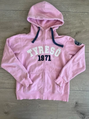 Rosa hoodie med tryck  - Cool rosa hoodie med huva och blå snören från Beachworks. Stort vitt och mörkblått tryck 'Tyresö 1971' på bröstet och patch på ena ärmen. Mjuk bomullskänsla och praktiska fickor framtill. Perfekt för chill dagar och att slänga på sig när det är lite kyligt. Den är i mycket bra skick!