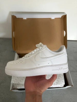 Nike Air Force 1  - Klassiska Nike Air Force 1 sneakers i helvitt läder med perforerad tåbox, vit sula och ikoniska swoosh-loggan på sidan. Skorna har lågt skaft och vita skosnören. Perfekt för dig som gillar stilrena och tidlösa sneakers.