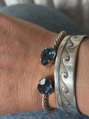 Caroline Svedbom armband med blå stenar - Säljer ett snyggt armband i silverfärg med tvinnad design och två stora blå stenar i ändarna. Armbandet är öppet och flexibelt, perfekt för att ge din outfit en cool touch. Passar dig som gillar stilrena men ändå unika smycken.har rostat lite inuti men annars jättebra titta på sista bild 