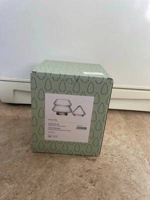 Glasburk formad som gran från Muurla - Säljer en glasburk från Muurla formad som en gran. Burken är transparent och har ett lock, perfekt för förvaring av godis eller småsaker. Förpackningen har ett grönt mönster med granar och tydlig produktbild på framsidan.