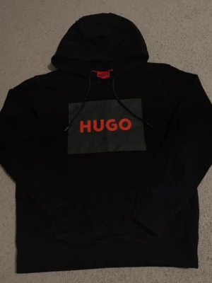 Hugo boss Hoodie - Säljer en svart hoodie från Hugo med stor röd logga på bröstet. Hoodien har huva med snören och ribbade muddar vid ärmslut och nederkant. Materialet är mjukt och bekvämt, perfekt för en avslappnad look.