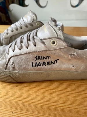 Saint Laurent vita låga sneakers - Vita låga sneakers från Saint Laurent med handskriven logga på sidan. Skorna har slitna detaljer, canvasmaterial och klassisk snörning. Perfekt för dig som gillar en edgy och avslappnad stil.
