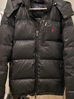 Svart dunjacka från Polo Ralph Lauren - Snygg svart glansig dunjacka från Polo Ralph Lauren med avtagbar huva och dragkedja framtill. Jackan har en röd logga broderad på bröstet, två sidofickor med dragkedja och en bröstficka. Perfekt för kalla vinterdagar och riktigt skön att bära.