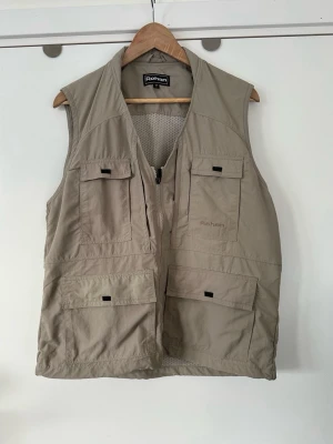 Beige utilityväst från Rohan - Säljer en beige utilityväst från Rohan med flera praktiska fickor framtill och meshfoder på insidan. Västen har dragkedja och är tillverkad i ett lätt och slitstarkt material, perfekt för dig som gillar funktionella plagg med stil.