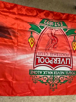 Liverpool FC röd supporterflagga - Stor röd supporterflagga med Liverpool FC:s emblem, texten 'You'll Never Walk Alone' och detaljer i grönt och gult. Tillverkad i tunt syntetmaterial, perfekt för att visa ditt stöd på matcher eller hemma.