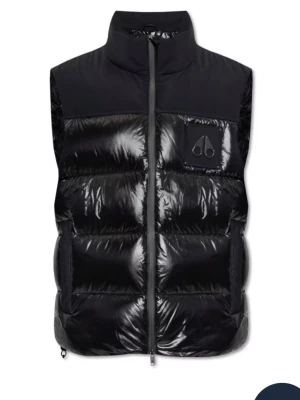 Svart dunväst från Moose Knuckles - Snygg svart dunväst från Moose Knuckles med glansig finish och hög krage. Västen har en cool logotyp på bröstet och är quiltad för extra värme. Perfekt att slänga över en hoodie eller t-shirt när det är kyligt ute.