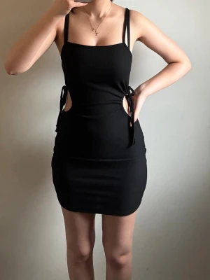 Svart ribbad kort klänning med snörning - Svart ribbad klänning med smala axelband och coola cut-outs på sidorna som knyts med snörning. Klänningen är tight och kort, perfekt för dig som gillar en edgy och trendig look. Materialet är mjukt och följsamt, vilket gör den bekväm att bära.