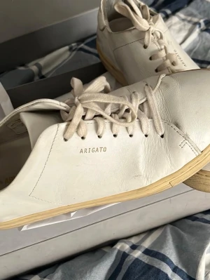 Vita sneakers från Axel Arigato - Snygga vita sneakers från Axel Arigato med minimalistisk design och diskret logotyp på sidan. Skorna är gjorda i läder och har vita snören samt en ljus beige sula. Perfekta för dig som gillar stilrena och trendiga skor. Säljes i originalkartong. Finns på Södermalm 