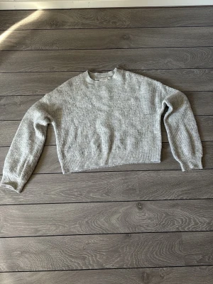 Grå stickad cropped tröja - En enkel och stilren grå stickad tröja med cropped passform och rund hals. Tröjan har långa ärmar och ribbstickade muddar vid ärmslut och nederkant. Perfekt för att matcha med jeans eller kjol.