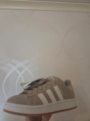 Beige Adidas Campus sneakers - Snygga beige Adidas Campus sneakers i mocka med klassiska vita ränder på sidorna och vit sula med gummidetalj. Låg modell med rund tå och snörning. Perfekta för en clean och stilren look. Färgen heter onebay. Har inte använts då de var för stora