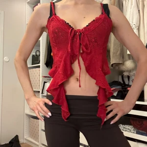 Röd spetsig omlott korsett-topp - Cool röd croppad topp med spetsdetaljer och omlott-design. Toppen har volangkant och snörning framtill som ger en edgy look. Ärmlös och croppad modell, perfekt för att sticka ut. Materialet känns som polyester och spets.