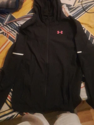 Svart vindjacka från Under Armour - Snygg svart vindjacka från Under Armour med huva och dragkedja framtill. Jackan har rosa logga på bröstet och reflekterande detaljer på ärmarna. Perfekt för blåsiga dagar och sportiga outfits.