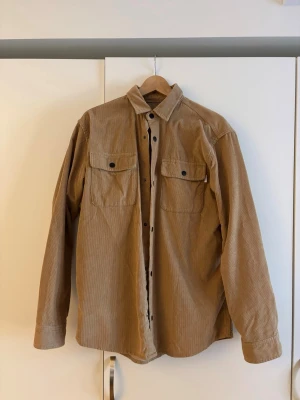 Beige vårjacka i manchester - Säljer en beige overshirt med klassisk krage och två bröstfickor med knappar. Overshirten har långa ärmar och är tillverkad i ett mjukt, strukturerat tyg som liknar bomull eller manchester. Perfekt att slänga över en t-shirt för en avslappnad look.