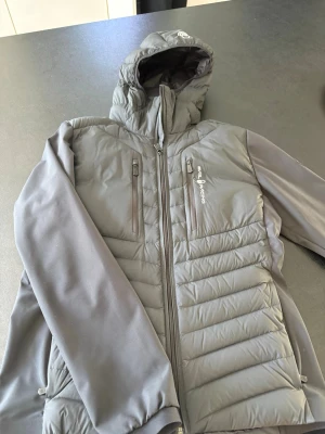 Grå/svart hybridjacka från Sail racing storlek M - Snygg grå hybridjacka från Sail racing med quiltad front och mjuka ärmar. Jackan har två bröstfickor med dragkedja, huva och logotyp på bröstet. Perfekt för lager-på-lager och outdoor. Materialet är en mix av syntet och softshell. Inga skador eller skavanker. Fräsch!