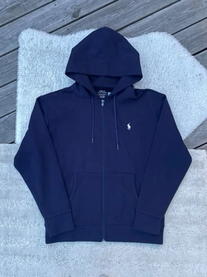 Mörkblå hoodie från Polo Ralph Lauren - Snygg mörkblå hoodie från Polo Ralph Lauren med dragkedja, huva och klassisk logga broderad på bröstet. Modellen har två fickor framtill och ribbade muddar. Perfekt för en chill och stilren look. Ny