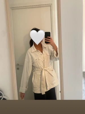 Stylish white denimjacket - Stilren vit denim jacka med knappar framtill, två bröstfickor och ett avtagbart knytskärp i midjan. Rak passform och klassisk krage ger en avslappnad men trendig look. Perfekt att slänga över en t-shirt för en cool lager-på-lager s