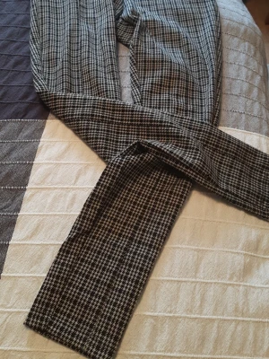 Kostymbyxor med hundtandsmönster - Snygga kostymbyxor med klassiskt svartvitt hundtandsmönster. Byxorna har raka ben och är tillverkade i ett mjukt och lätt material som ger en stilren look. Perfekta för dig som vill ha en trendig och tidlös stil.