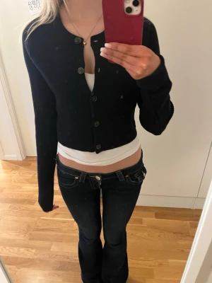 Mörkblå cropped kofta med knappar - Snygg mörkblå cropped kofta med långa ärmar och knäppning framtill. Klassisk rund halsringning och ribbstickad struktur. Perfekt att styla över ett linne eller t-shirt för en trendig lager-på-lager-look.