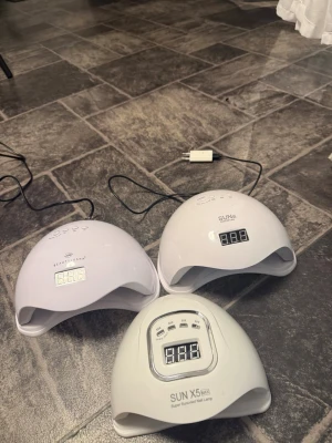 Tre vita UV/LED nagellampor - Tre vita nagellampor för UV/LED-gel, med digital display och flera timerinställningar. Perfekta för att härda gel- och shellacklack. Märkena är SUN, SUN X5 MAX och BEAUTY & SUNSHY. Alla lampor har rundad form och är tillverkade i plast.