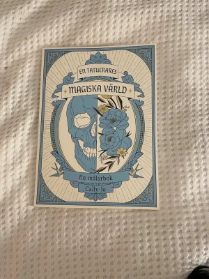 En tatuerarens magiska värld målarbok - En kreativ målarbok med tatueringstema av Cally-Jo. Omslaget har en blå och vit design med en dödskalle och blommor, samt dekorativa detaljer. Baksidan pryds av fåglar och blommor i blått och gult. Perfekt för dig som gillar konst och tatueringar.