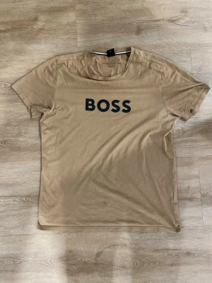 Hugo Boss T-Shirt  - Säljer en Hugo Boss T-Shirt i beige/brun färg. Mycket bra kvalitet som vanligt. Tröjan har en liten fläck på framsidan som man inte ser om man inte kollar noggrant. Vid frågar är det bara att ställa de ☺️