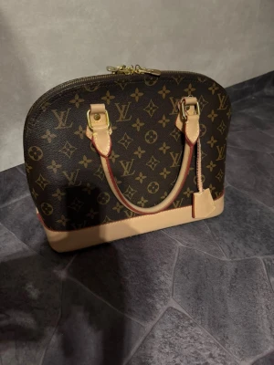  handväska  - Säljer en klassisk handväska Alma handväska i brunt monogrammönster med beigea läderdetaljer och guldfärgade metalldelar. Väskan har två handtag och en stabil botten med metallfötter. Perfekt för dig som gillar lyxiga accessoarer.